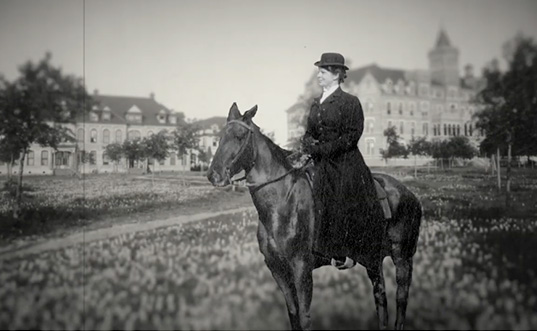 A woman dressed in a black dress and large hat sits atop a horse in front of TCU buildings